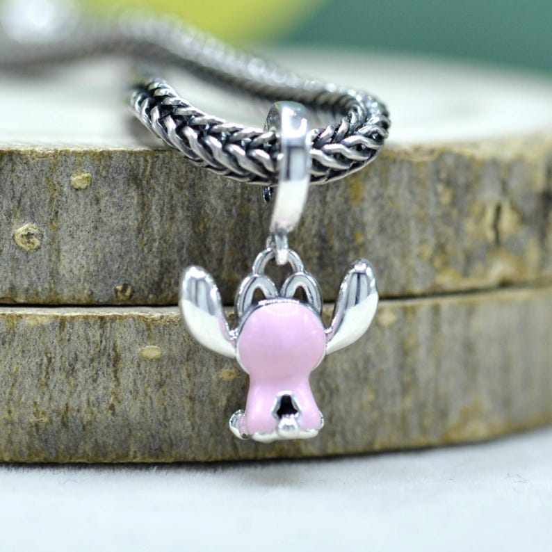 May include: A silver charm pendant featuring a pink cartoon character with outstretched arms. The charm is attached to a silver chain. The pendant is set against a wooden background.