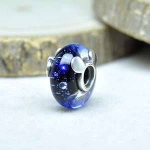 May include: A blue and black glass bead with a galaxy-like design. The bead has silver-colored metal accents and a hole for stringing. The bead is oval-shaped and has a shiny, reflective surface.