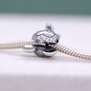 May include: A silver charm with a floral design, encrusted with small, sparkling crystals. The charm is attached to a silver chain, and features a small, round accent on top. The charm is designed to be a decorative element for a bracelet or necklace.