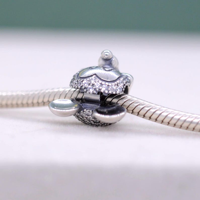 May include: A silver charm with a floral design, encrusted with small, sparkling crystals. The charm is attached to a silver chain, and features a small, round accent on top. The charm is designed to be a decorative element for a bracelet or necklace.