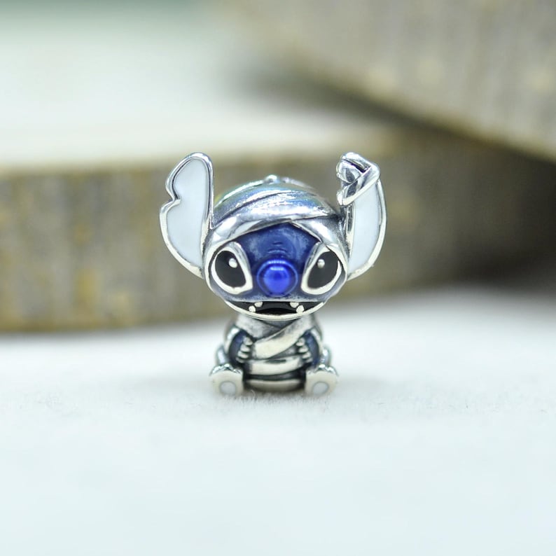 Może przedstawiać: Srebrny koralik typu charms w kształcie postaci Stitcha owiniętego w bandaże. Wz&oacute;r charakteryzuje się głębokim niebieskim emaliowaniem na głowie i ciele, białym emaliowaniem na dużych uszach oraz małym niebieskim nosem. Metal ma polerowane wykończenie.