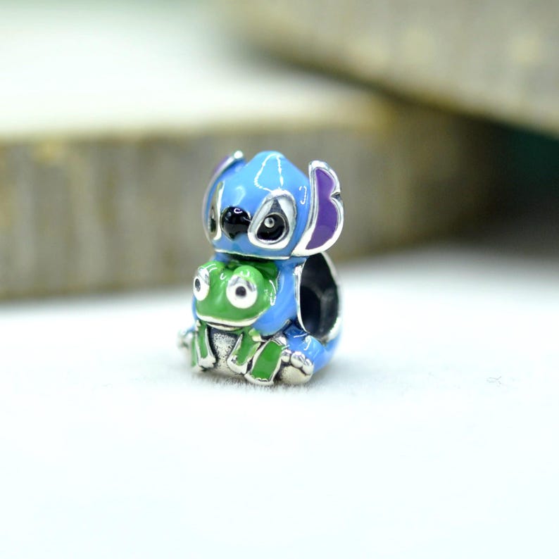 May include: A silver-toned metal charm bead featuring a blue character with large ears and a black nose, holding a small green frog. The bead has a hollow center for threading onto a bracelet, with vibrant enamel detailing in blue, green, and purple.