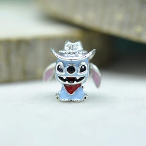 May include: A silver-toned metal charm featuring a light blue character with large black eyes, a wide grin, and pink inner ears. The character wears a silver cowboy hat and a small red bandana around its neck, crafted with detailed enamel accents.
