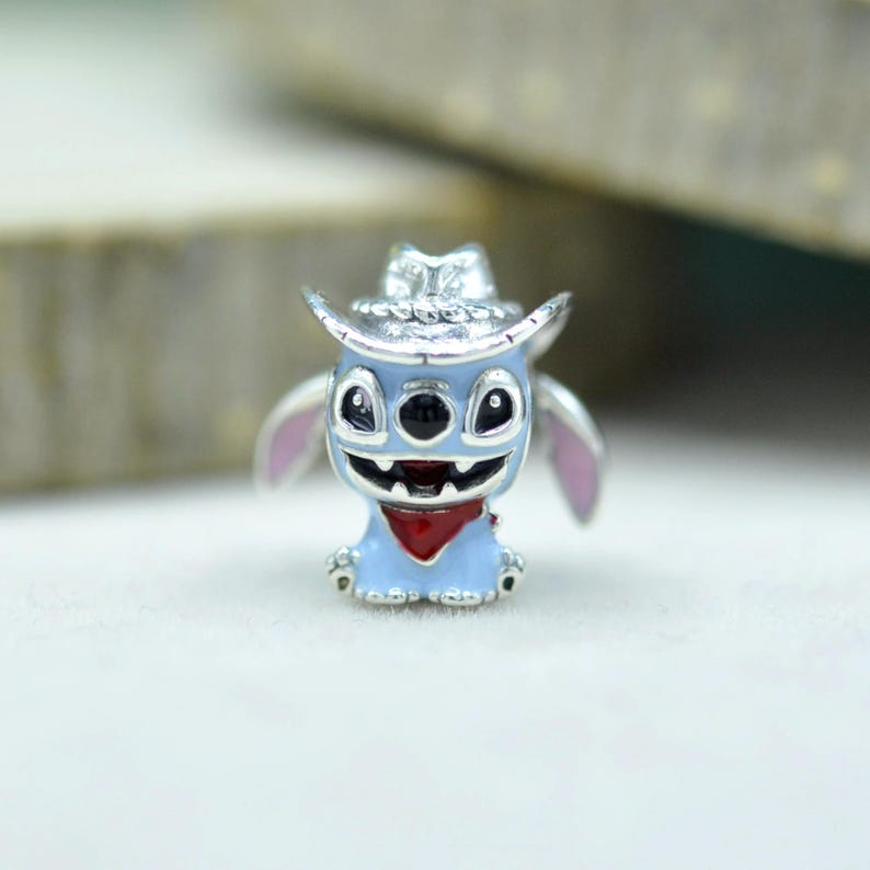May include: A silver-toned metal charm featuring a light blue character with large black eyes, a wide grin, and pink inner ears. The character wears a silver cowboy hat and a small red bandana around its neck, crafted with detailed enamel accents.