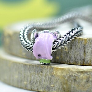 May include: A silver charm bead featuring a light pink enameled character figure with a small green base, threaded onto a braided silver chain. The side of the charm is engraved with the text Disney and Pandora in a clear, legible font.