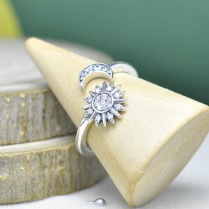 以下が含まれることがあります： 太陽と月のデザインが施されたシルバーリング。太陽は、中心にクリアストーンのクラスターと放射状の光線があります。三日月には、小さなクリアストーンが飾られています。リングは木の円錐の上に展示されています。