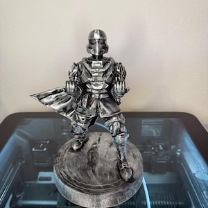 May include: A silver-colored figurine of a futuristic armored soldier standing on a circular base. The character wears a helmet with a visor, chest plate, and a flowing cape, with both hands held forward in a clenched fist position.
