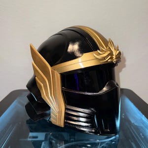 May include: A black and gold helmet with a glossy finish. The helmet features gold accents, including a band across the top and stylized wing-like details on the sides. The design is reminiscent of a superhero or fantasy character.