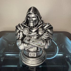 May include: A silver-colored bust of a masked figure wearing a hooded cloak and armor with arms crossed. The figure features a metallic finish with dark shading in the crevices, a prominent face mask, and circular emblems on the chest area.