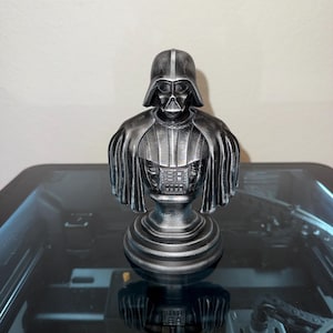 Puede incluir: Busto gris metálico de Darth Vader, personaje de Star Wars. El busto presenta una representación detallada del casco, la máscara y la armadura de Vader. La escultura está montada sobre una base circular.