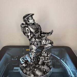 May include: A silver-colored figurine of a soldier in futuristic armor, kneeling on a textured base. The figure has a helmet with a visor and is posed with both hands extended forward as if holding an object. The finish has a metallic sheen.