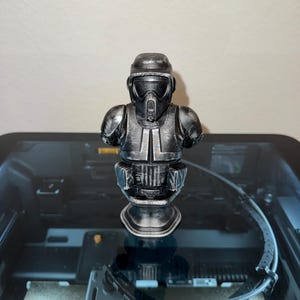 May include: A metallic gray bust of a Star Wars Scout Trooper, featuring a helmet and armor. The figure is displayed on a dark surface, showcasing the details of the design. The bust is a collectible item.