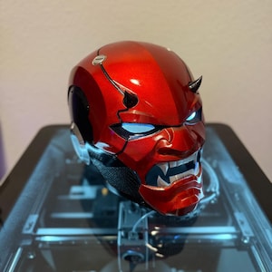 May include: A red and black helmet with a glossy finish, featuring a demonic face design with glowing blue eyes and small horns. The helmet is displayed on a clear surface.