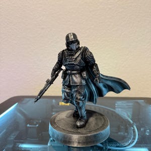 May include: A black figurine of a character in armor, holding a rifle, and standing on a circular base. The figure has a helmet and a cape. The figurine is likely a collectible or a model.