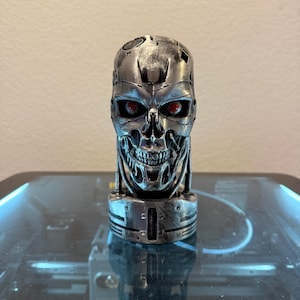 May include: A metallic silver robotic skull sculpture featuring a weathered, distressed finish with intricate mechanical details, exposed wiring, and two glowing red eyes set within deep sockets, mounted on a cylindrical base with vertical grooves.