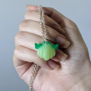 Pode incluir: Colar de corrente prateada com um pingente verde claro em forma de ab&oacute;bora estilizada com detalhes verdes em forma de asa. O pingente &eacute; segurado em uma m&atilde;o contra um fundo neutro.