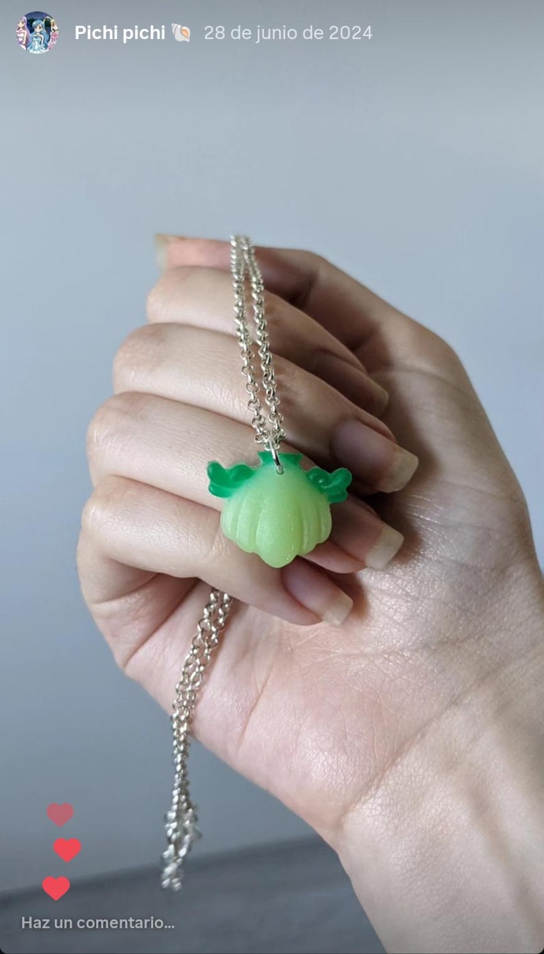 Pode incluir: Colar de corrente prateada com um pingente verde claro em forma de ab&oacute;bora estilizada com detalhes verdes em forma de asa. O pingente &eacute; segurado em uma m&atilde;o contra um fundo neutro.