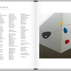 May include: A mobile sculpture with colorful, abstract shapes hanging from thin wires. The shapes are in shades of red, orange, yellow, blue, and purple. The mobile is suspended from the ceiling of a white room.