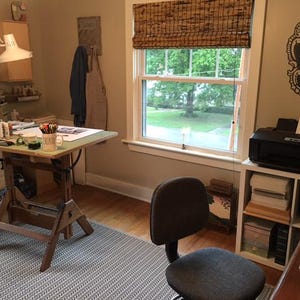 Pode incluir: Um est&uacute;dio de arte bem iluminado com uma mesa de desenho de madeira, uma l&acirc;mpada de mesa branca e uma impressora. A sala tem uma janela com uma cortina tecida, um tapete cinza estampado e uma cadeira de escrit&oacute;rio preta. Materiais de arte s&atilde;o vis&iacute;veis.