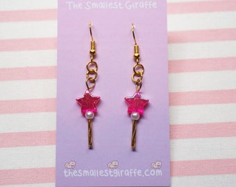 May include: A pair of gold dangle earrings with pink star-shaped charms and white pearl beads. The earrings are on a pink card with the text "The Smallest Giraffe" and "thesmallestgiraffe.com".
