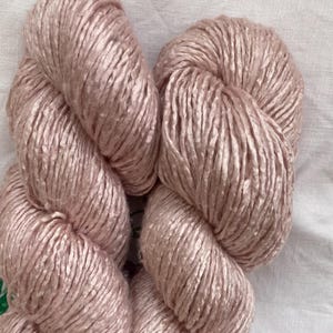 May include: Two skeins of blush pink yarn are displayed against a white background. The yarn is tightly wound, showing a textured surface. The color is a soft, muted pink, ideal for knitting or crochet projects.