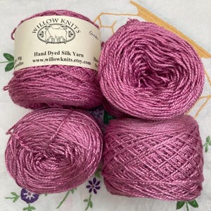 May include: Four skeins of shiny, magenta-coloured hand-dyed silk yarn. One skein features a paper label with the text Willow Knits, Hand Dyed Silk Yarn, and www.willowknits.etsy.com, along with a graphic of a sheep. The yarn has a smooth texture.