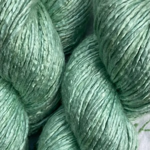 May include: Close-up of several skeins of mint green yarn. The yarn has a soft, textured appearance, with visible strands and a slight sheen. The color is a muted, pastel green, ideal for knitting or crochet projects.