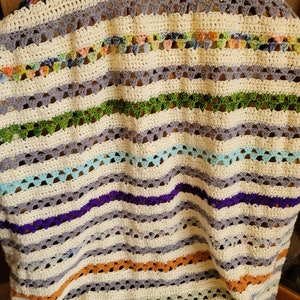 Puede incluir: Una manta de ganchillo con un patrón de rayas en varios colores, incluyendo verde, azul, morado, naranja y blanco. La manta está colocada sobre una silla de madera.