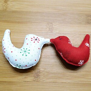 May include: Two fabric bird ornaments. One is white with red and green polka dots and snowflake designs. The other is red with white snowflake designs. The birds are positioned as if they are kissing.