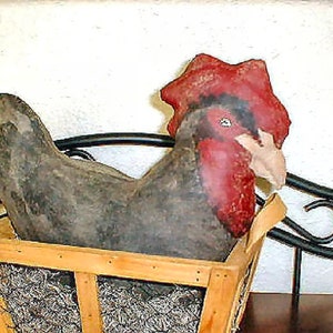 Puede incluir: Una figura de gallo negro y rojo sentada en una cesta de madera con paja. El gallo tiene una cresta y una papada rojas y un cuerpo negro.