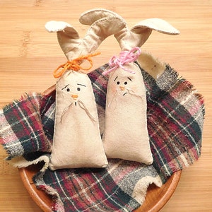 May include: Two handmade fabric bunny rabbits with embroidered faces. The bunnies are sitting on a plaid fabric in a wooden bowl. One bunny has a pink bow and the other has an orange bow.