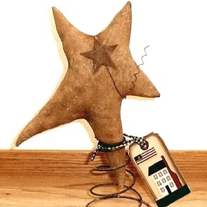 Könnte beinhalten: Ein brauner Stoffstern mit einem kleineren braunen Stern oben, der an einer Metallfeder befestigt ist. Der Stern ist mit einem schwarz-weiß karierten Band und einem Etikett mit einer rot-weiß-blauen amerikanischen Flagge und einer Haus-Illustration gebunden.