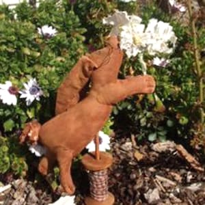 Peut inclure: Une figurine de lapin en tissu brun, faite à la main, avec une bobine de fil. Le lapin est debout sur un lit de paillis brun et de plantes vertes. Le lapin tient une fleur blanche dans sa main.