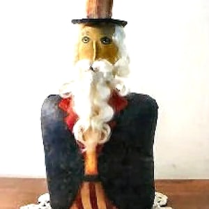 May include: A handmade doll figurine of a person wearing a red, white and blue striped suit, a black top hat, and a white beard. The doll is standing on a white crocheted doily.