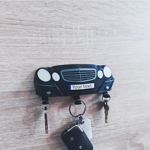 May include: A black car-shaped key holder with the text "Your text" on a license plate. The key holder has three key rings with keys attached. The key holder is mounted on a light wood-grain surface.