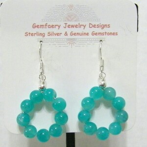 May include: A pair of turquoise gemstone earrings. Each earring features a circular design made of turquoise beads, suspended from a silver hook. The earrings are displayed on a white card with text that reads "Gemfaery Jewelry Designs Sterling Silver & Genuine Gemstones."
