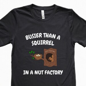 May include: Black t-shirt with the text "BUSIER THAN A SQUIRREL IN A NUT FACTORY" in white. The design includes a cartoon squirrel in a tree trunk and a nest with eggs.