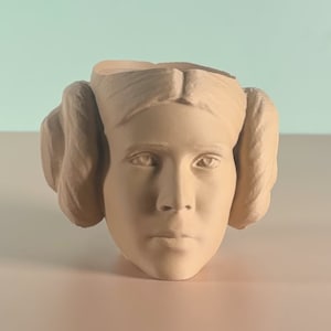 Può includere: Un vaso di fiori in ceramica beige chiaro a forma di testa di donna con chignon laterali. Il vaso ha un viso dettagliato con gli occhi chiusi e un'espressione neutra. La parte superiore della testa è aperta per la piantagione.