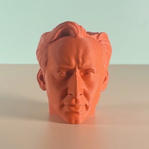 以下が含まれることがあります： オレンジ色の3Dプリントされた男性の頭部の胸像。胸像は詳細で現実的で、眉間にしわを寄せ、真剣な表情をしている。