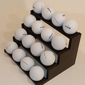 May include: A black tiered stand holds a collection of white golf balls. The balls are arranged in rows, with the top row having four balls, the middle row having five balls, and the bottom row having six balls. The balls are all marked with the word "Wilson" and a number.