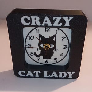 May include: Black and white clock with a black cat design. The clock face says "Crazy Cat Lady".