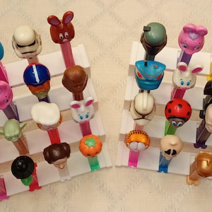 May include: A collection of 24 colorful plastic figurines on white stands. The figurines are arranged in two rows of 12. Each figurine is a different character, including a bunny, a turtle, a ladybug, a robot, a pumpkin, and a cat.