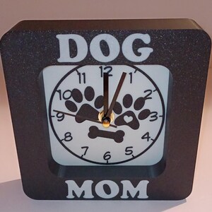 May include: Black square clock with white lettering that says "DOG MOM" and a white clock face with black paw prints and a bone. The clock hands are black.