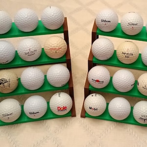 May include: A green golf ball rack with four shelves, holding a variety of golf balls. The rack is filled with white golf balls with different brand names and logos, including Top-Flite, Wilson, Titleist, and others.