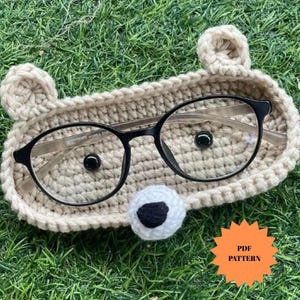 Puede incluir: Un estuche para gafas de crochet beige con forma de oso, con gafas de montura negra en su interior. El soporte tiene dos orejas pequeñas y una nariz blanca y negra. Un gráfico de patrón PDF está en la esquina inferior derecha.