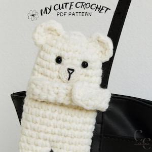Könnte beinhalten: Ein weißer gehäkelter Bär mit schwarzen Augen und einer schwarzen Nase ist in diesem Bild zu sehen. Der Bär hält eine schwarze Tasche fest. Der Text "MY CUTE CROCHET PDF PATTERN" befindet sich oben im Bild.