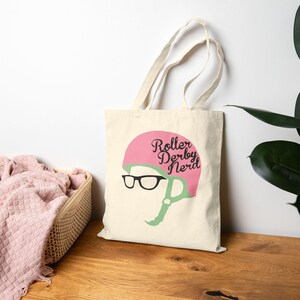 Könnte beinhalten: Cremefarbene Tragetasche mit einem rosa Roller Derby Helm-Design und den Worten "Roller Derby Nerd" in schwarzer Schrift. Der Helm hat einen grünen Kinnriemen und eine schwarze Brille. Die Tasche hat zwei cremefarbene Griffe.
