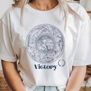 May include: White t-shirt with a blue and white graphic of a circular design with a figure in the center. The text "Victory" is printed below the graphic with a laurel wreath.