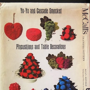 May include: A vintage McCall's transfer pattern for making yo-yo and cascade smocked pincushions and table decorations. The pattern includes instructions for making a heart-shaped pincushion, a round pincushion, a grape cluster, a cluster of red balls, a cluster of red balls on a plate, a cluster of red and purple balls, a red flower in a pot, a purple flower in a pot, and a blue flower in a pot. The pattern is for McCall's Multi-Blue 6988.