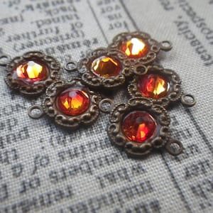 May include: A collection of bronze-toned connectors, each featuring a central, faceted, orange-red gemstone. The connectors have a decorative, floral-inspired frame and small loops. The gemstones display a range of fiery hues.